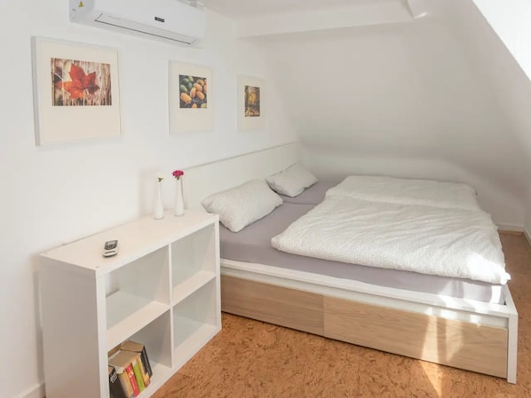 A small, modern bedroom with a loft-style bed, white walls, and a wooden floor. There are three framed pictures on the wall, a small white console table, and a bookshelf.