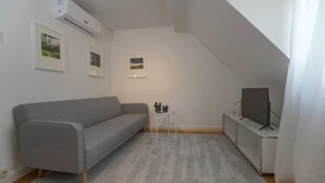 Un petit salon moderne avec un canapé gris, une petite table agrémentée d’objets décoratifs, un téléviseur posé sur un meuble et des tableaux encadrés accrochés au mur.