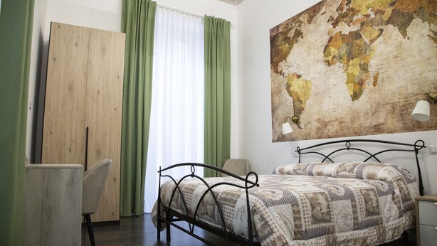 Een slaapkamer met een bed, een bureau, een stoel, een kaart aan de muur en een raam met gordijnen.
