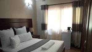 A hotel room with a large bed, a bedside table with a lamp, a small desk with a coffee maker, and curtains.