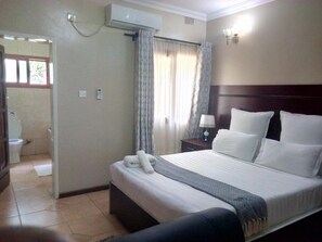 A hotel room with a bed, a nightstand, a lamp, a window with curtains, an air conditioning unit, and a bathroom visible through an open door. - Zenith lodge (Lusaka)
