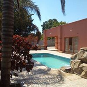 A modern building with a swimming pool, surrounded by palm trees and a rock feature. - Zenith lodge (Lusaka)