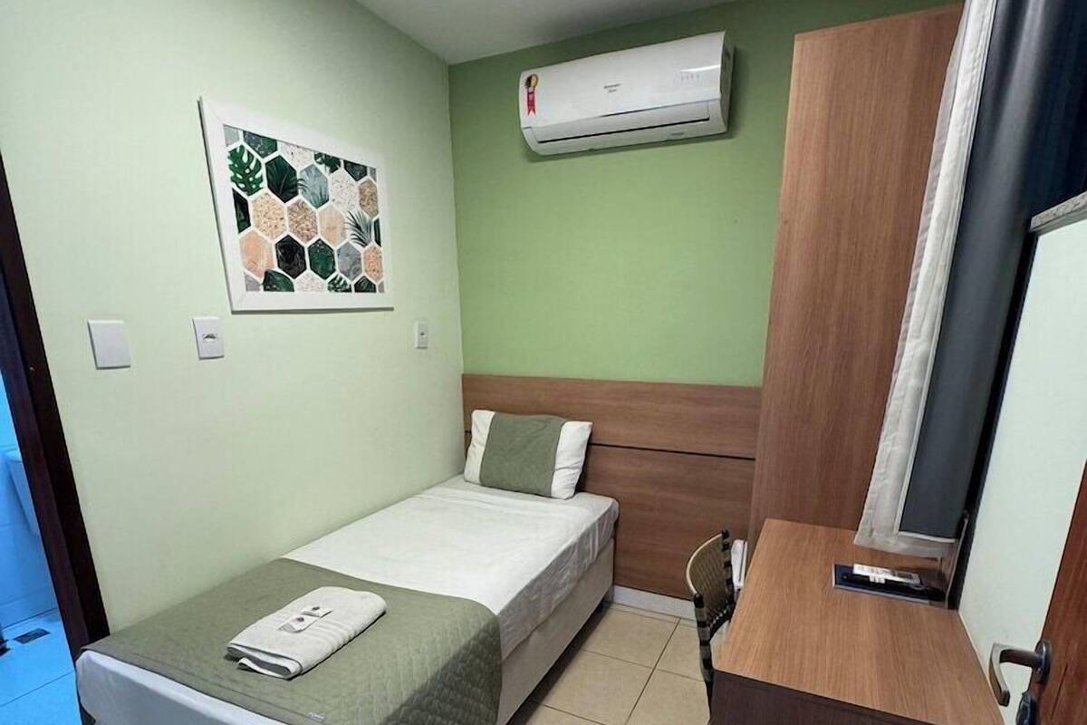 A small, clean room with a bed, a desk, an air conditioning unit, and a wall-mounted artwork.