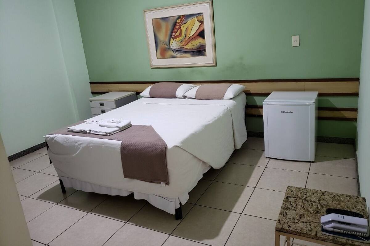 A room with a bed, a small bedside table, a nightstand, a refrigerator, and a painting on the wall.