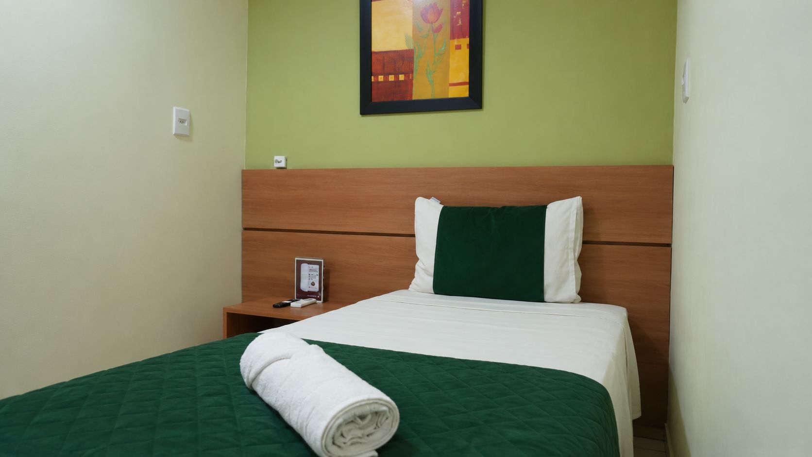 A hotel room with a bed, a nightstand, a painting on the wall, and a green wall.
