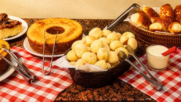 A buffet table with a variety of food items including a cake, bread rolls, and a bowl of yellow pastries, possibly cheese curds, with serving utensils. - Domus Hotel Veneza - Ipatinga (Ipatinga)