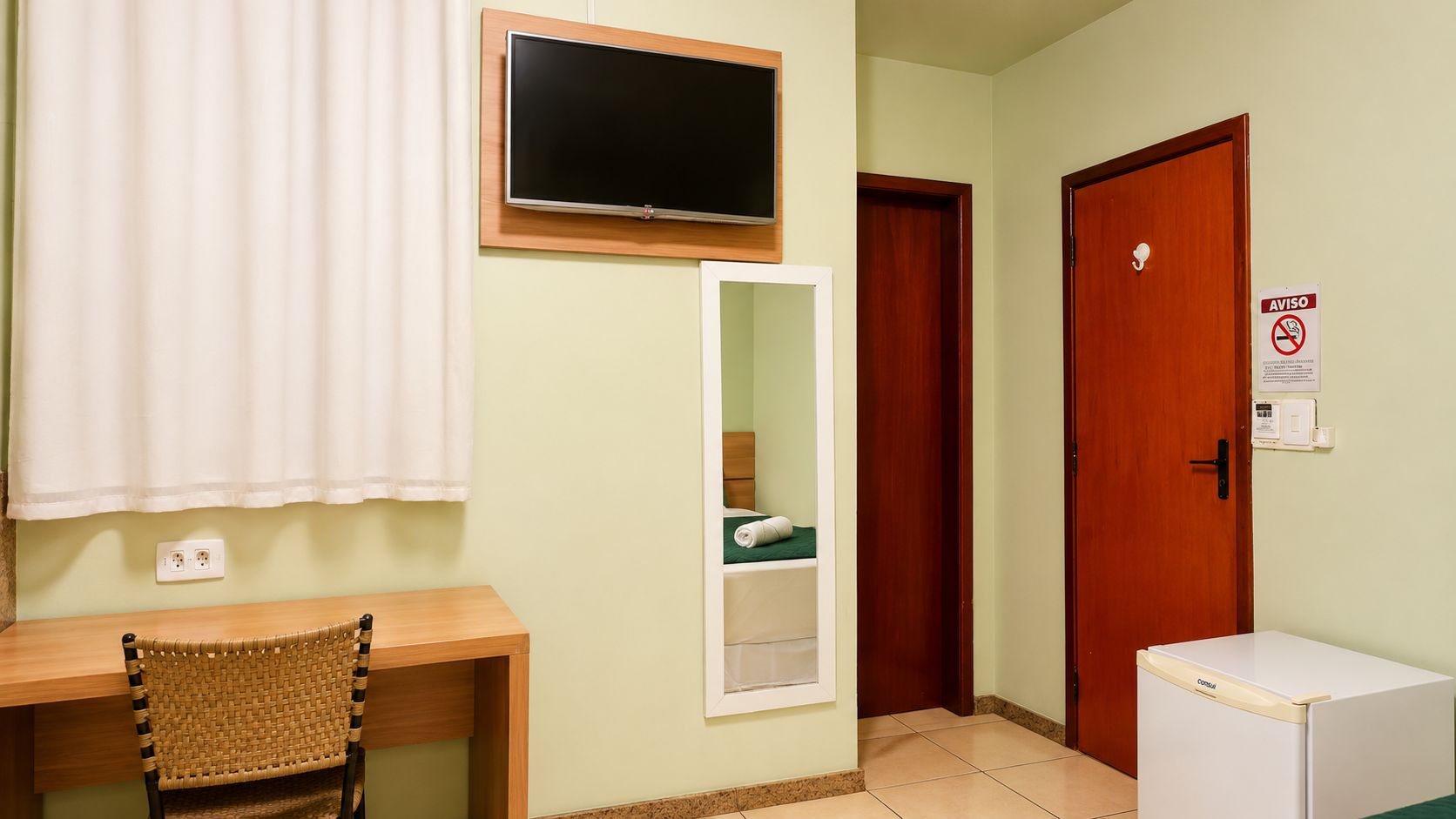 Ein Zimmer mit einem Holztisch, einem darüber montierten Fernseher, einem Spiegel, einer Tür mit einem AVISO-Schild und einem kleinen Kühlschrank.
