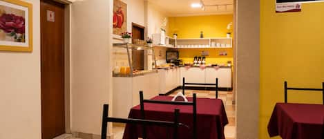 Un comedor con mesas dispuestas para los huéspedes, una cocina al fondo y un letrero que indica una sala de usos múltiples.