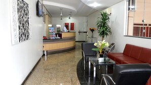 Zona de recepción con mostrador, un sofá rojo, una mesa de vidrio con un florero y una planta grande.