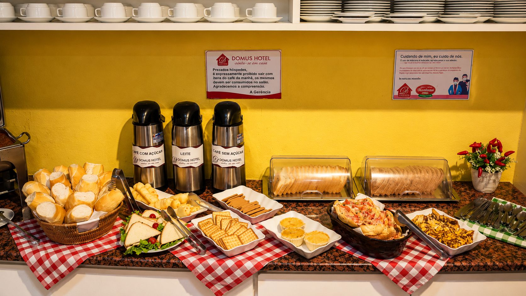 Disposição de buffet com diversos alimentos, incluindo pães, sanduíches e doces, sobre uma toalha xadrez.
