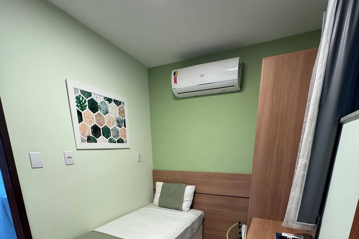 A small, clean room with a bed, a wooden cabinet, a wall-mounted air conditioner, and a framed botanical print.