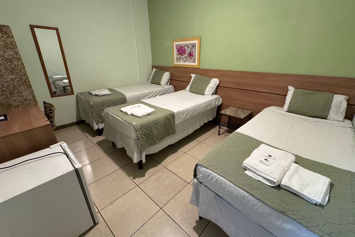 A hotel room with two single beds, a larger bed, a wooden headboard, a small table, a mirror, and a framed floral artwork on the wall.
