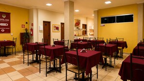 Uma sala de jantar de hotel com várias mesas e cadeiras, uma receção e uma cozinha ao fundo.