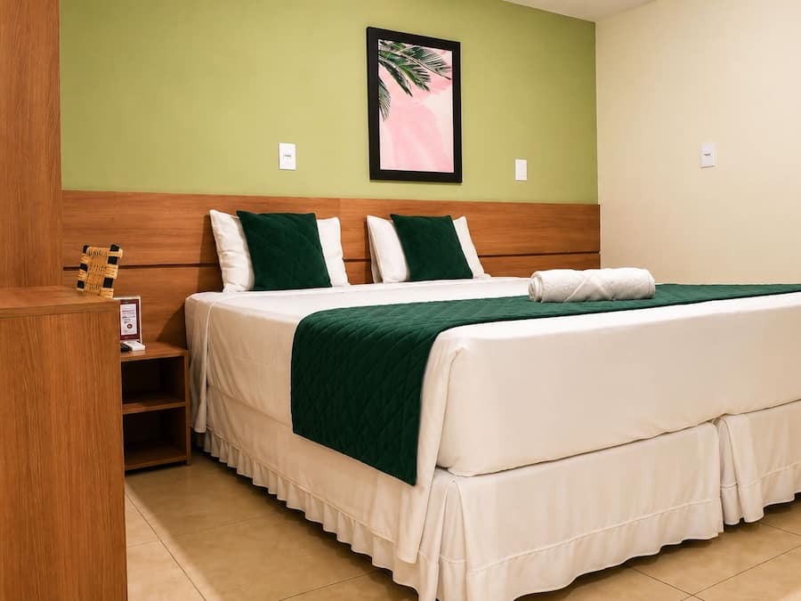 A hotel room with a wooden headboard, a bed with white bedding and green throw pillows, a small wooden nightstand, and a framed picture on the wall.
