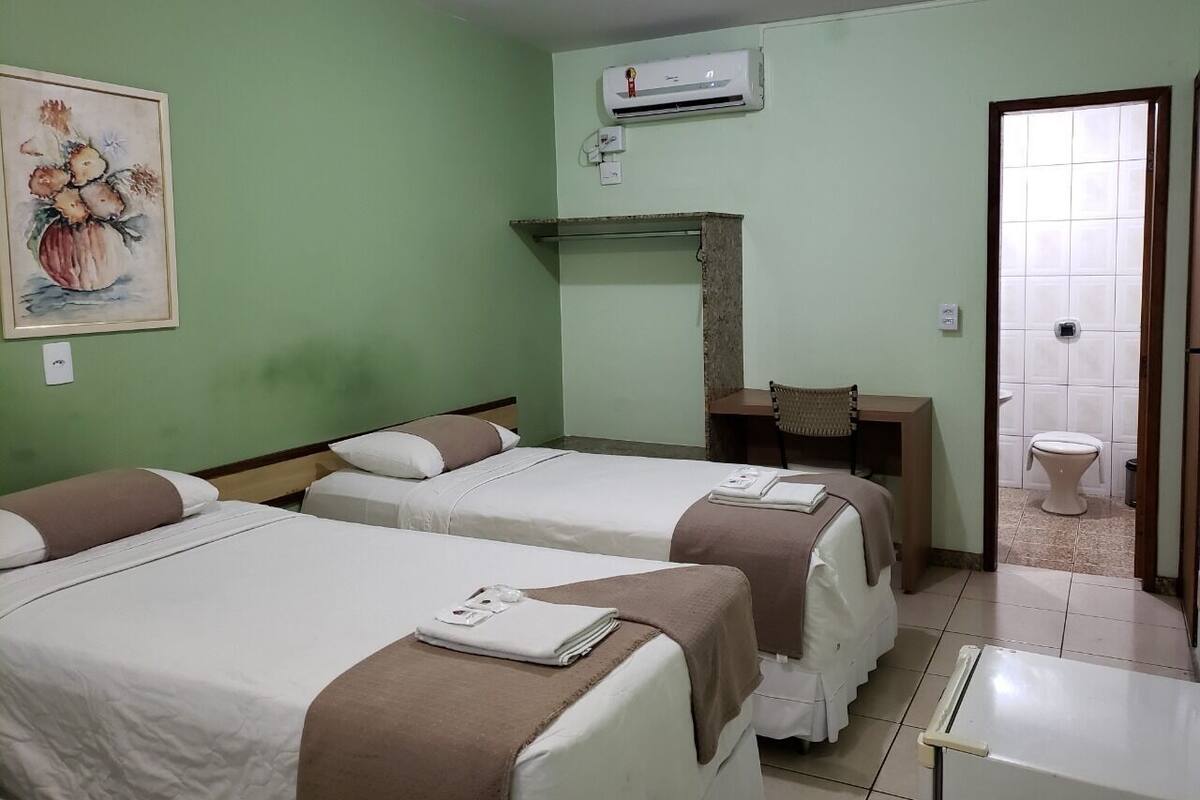 A hotel room with three beds, a desk, a chair, an air conditioning unit, a painting, and a bathroom visible through an open door.