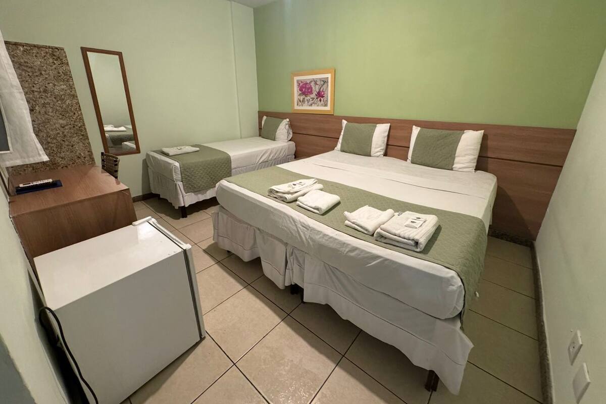 A hotel room with a large bed, two smaller beds, a desk with a chair, a mirror, and a small refrigerator.