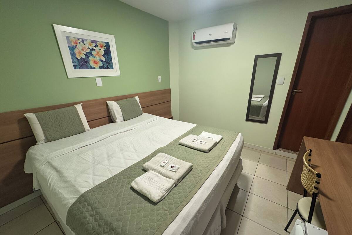 A neatly made bed with a green quilt, a framed floral artwork on the wall, and a mirror above the door.