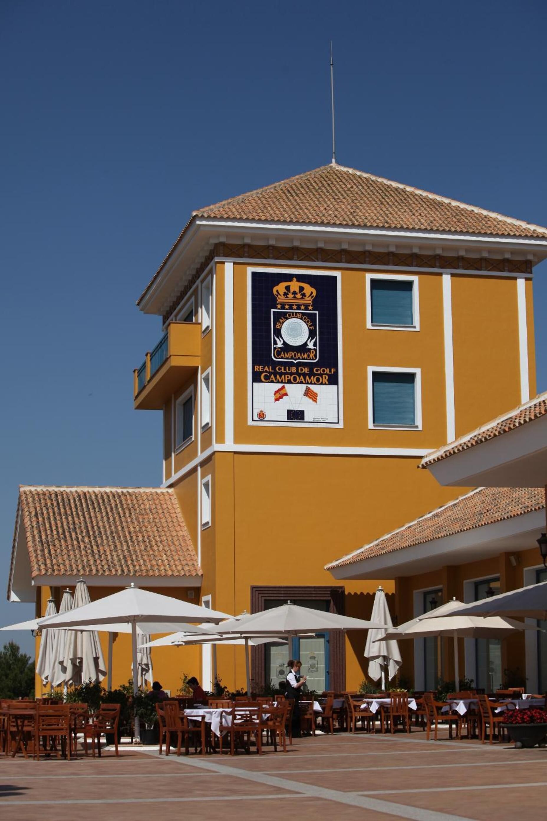 Un bâtiment jaune avec une enseigne indiquant un club de golf, des tables à l’extérieur et des parasols.