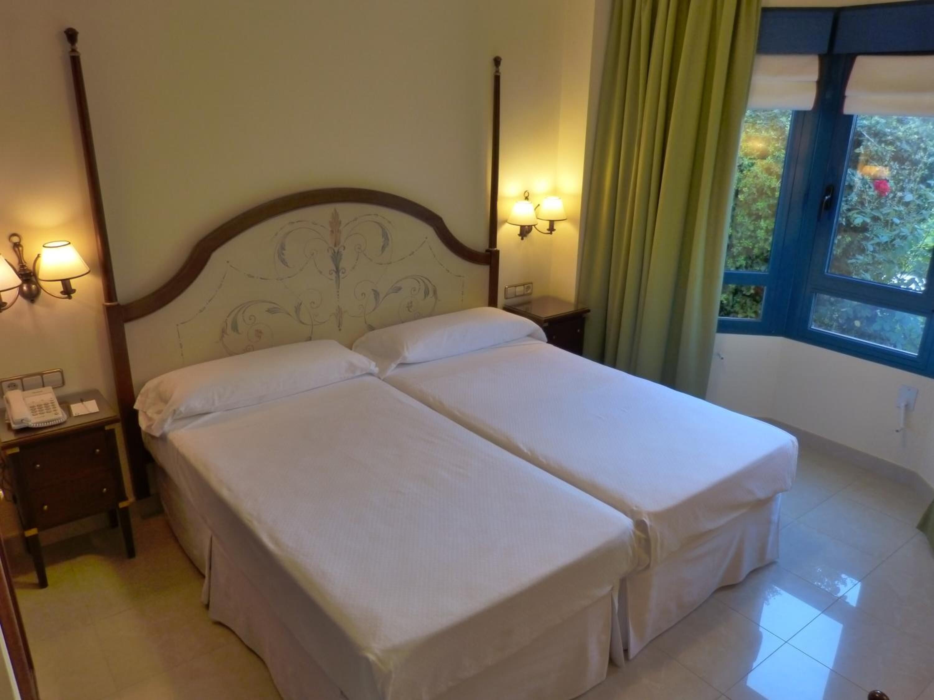 A double bed with a wooden headboard, two bedside tables with lamps, a telephone, and a window with curtains.
