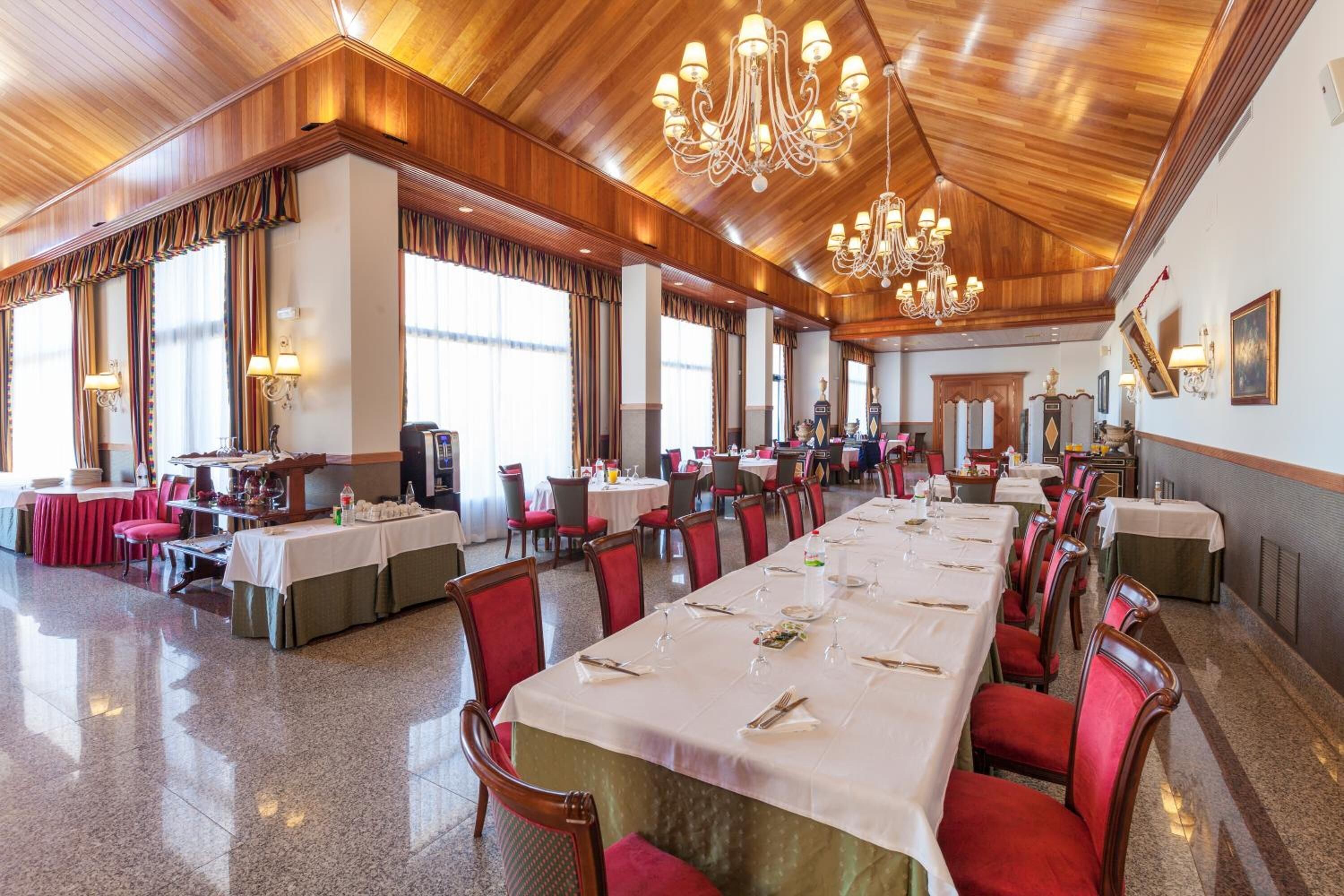 A spacious banquet hall with long tables set for dining, large windows, and a high ceiling with chandeliers.