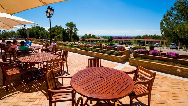 Un patio al aire libre con mesas y sillas de madera, rodeado de macizos de flores y vistas a un estacionamiento y un paisaje lejano.