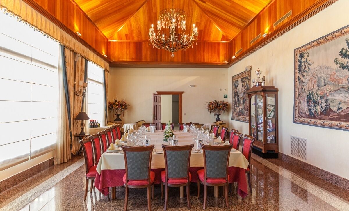 A formal dining room with a long table set for a meal, wooden paneling on the ceiling, and a large tapestry on the wall.