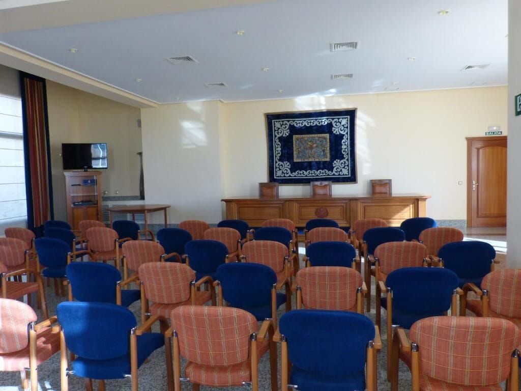 Un salón de conferencias con hileras de sillas, un televisor sobre un pedestal y un tapiz decorativo.