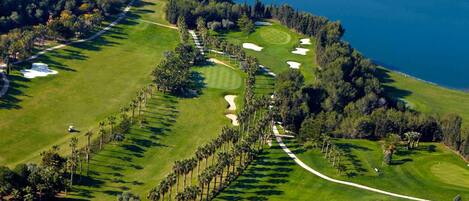 An aerial view of a golf course with sand bunkers, palm trees, and a coastal backdrop.
