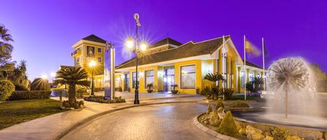 A hotel with a well-lit entrance, a fountain, and a clear sky.