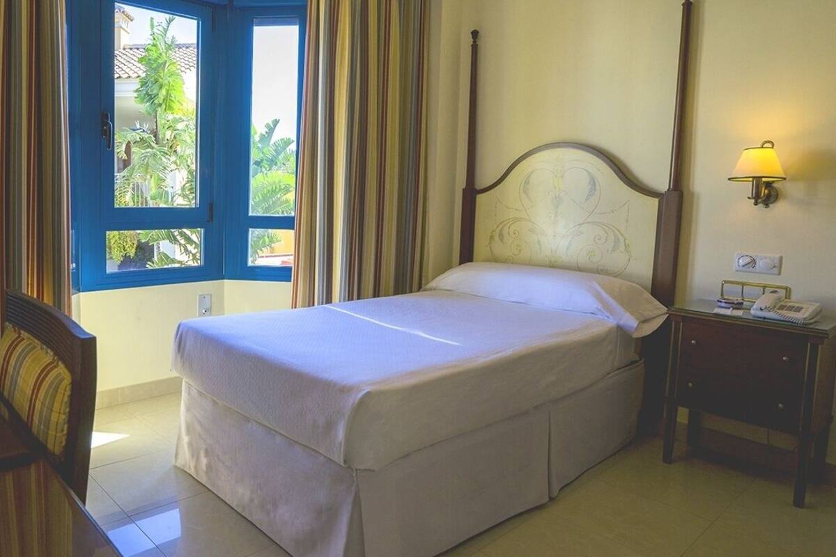 Une chambre d’hôtel avec un lit, un bureau avec une lampe, une table de chevet avec un téléphone et une fenêtre avec des rideaux.