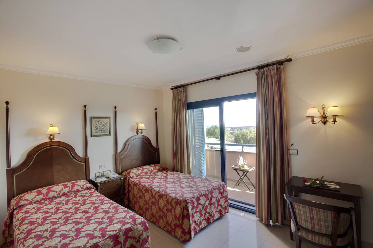 Une chambre d’hôtel avec deux lits, une petite table, une chaise et un balcon avec des rideaux.