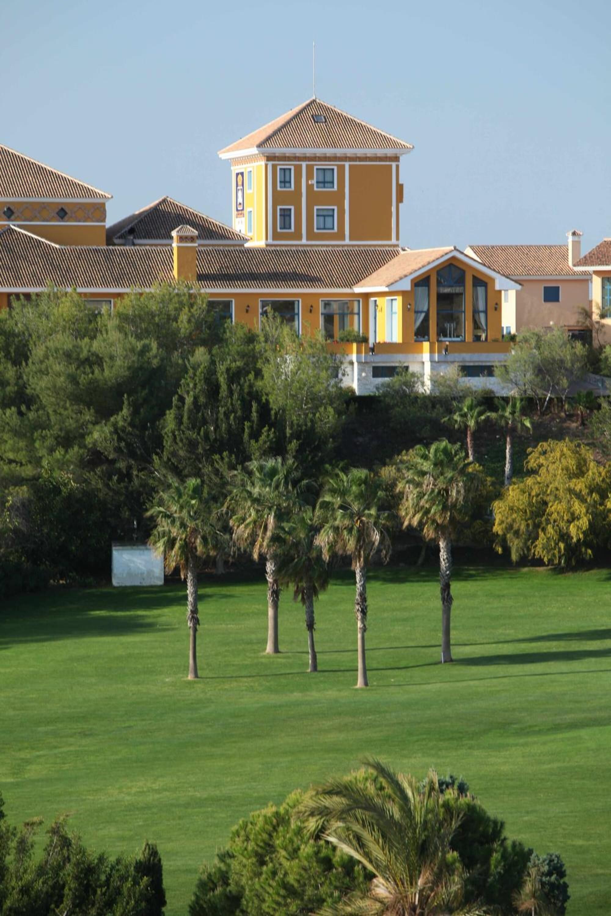 Un gran edificio amarillo con una torre destacada, rodeado de exuberante vegetación y palmeras.
