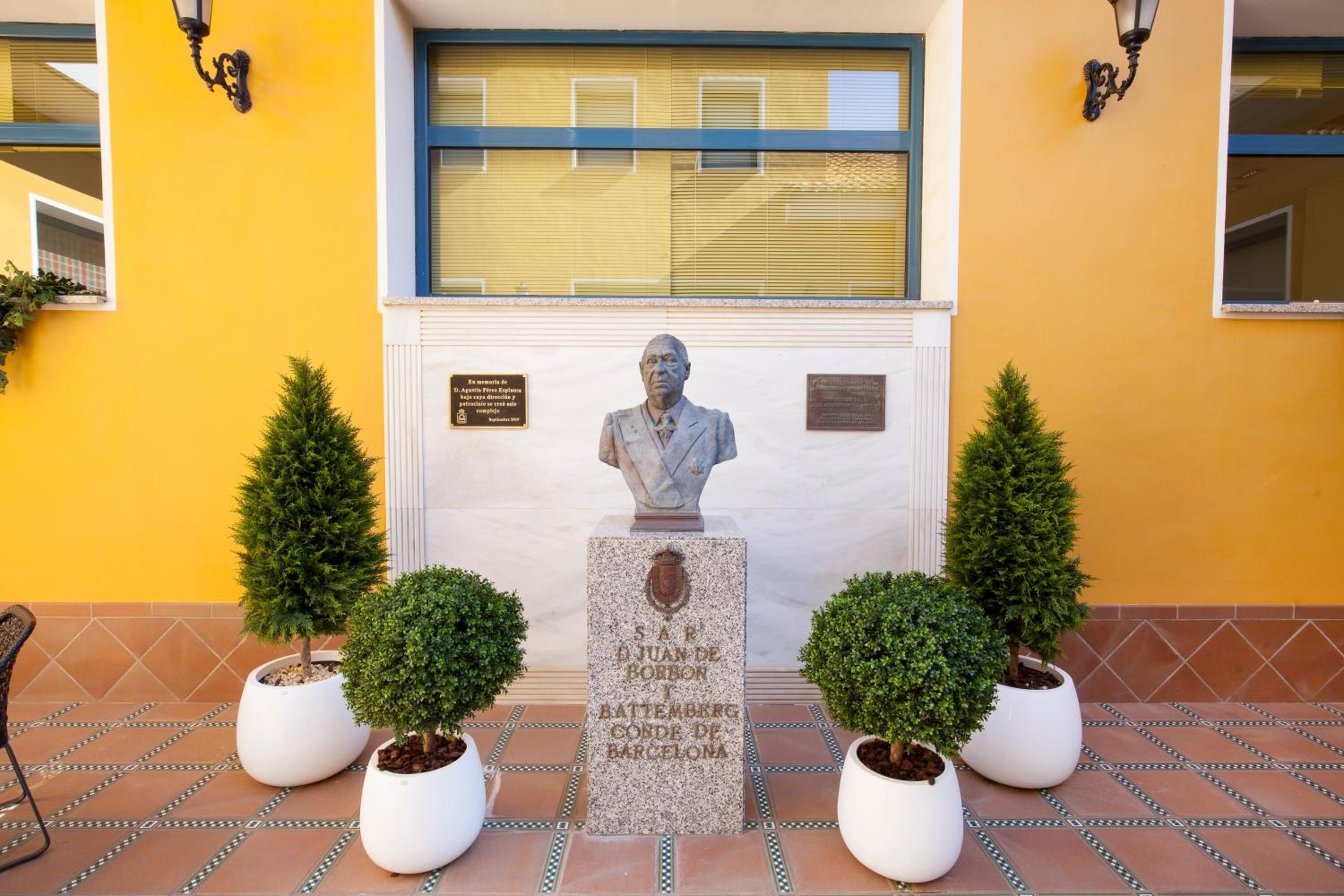 Eine Statue eines Mannes im Anzug, umgeben von Topfpflanzen, mit einer Tafel davor.