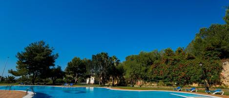 A clear blue swimming pool with a wooden deck, surrounded by trees and a few lounge chairs.