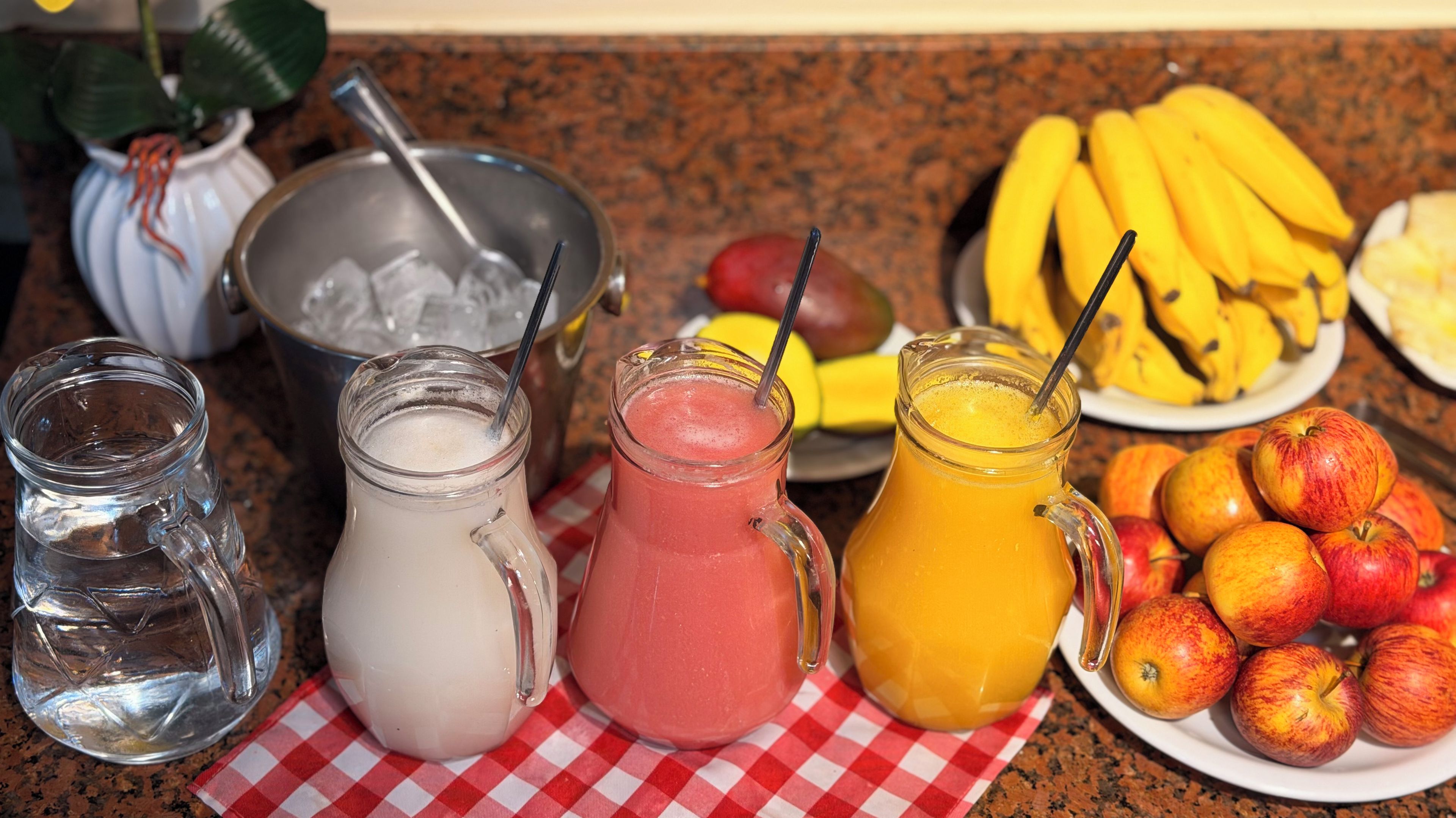 Een tafel met drie glazen karafjes met verschillende soorten drankjes, een schaal ijs, een bord bananen en een bord appels.