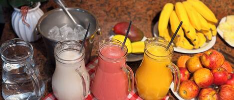 Een tafel met drie glazen karafjes met verschillende soorten drankjes, een schaal ijs, een bord bananen en een bord appels.