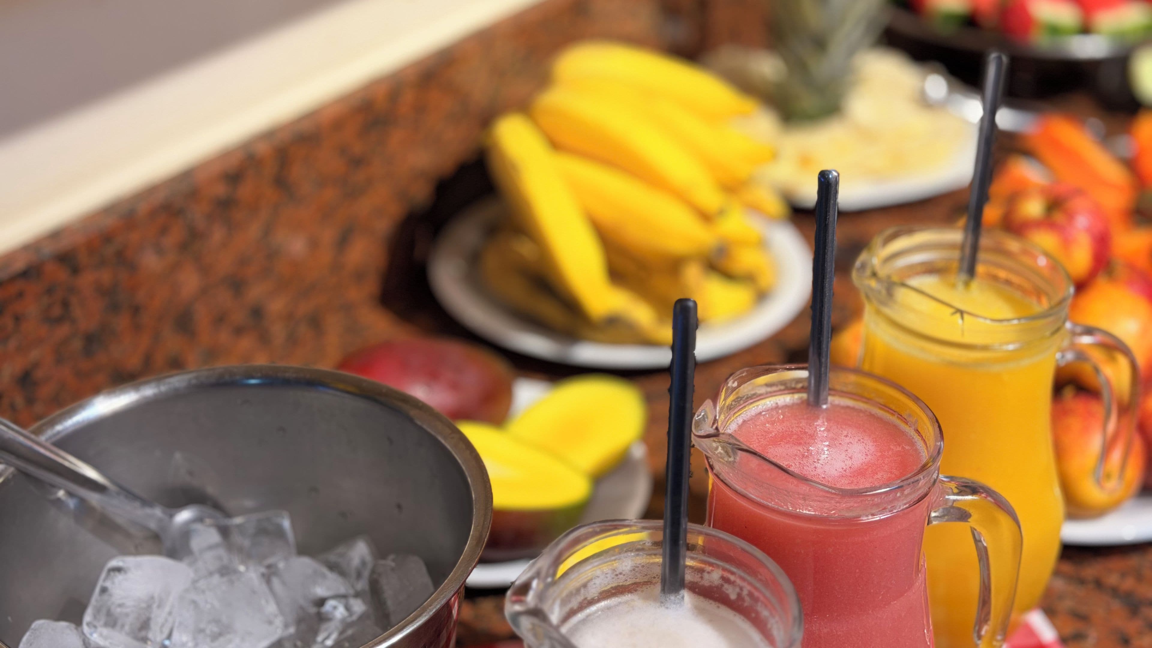 Una mesa de buffet con diversas frutas, un bol de hielo y dos zumos de fruta con pajitas.