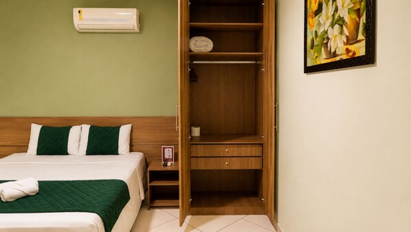 A hotel room with a bed, a wooden wardrobe, a painting on the wall, and an air conditioning unit.