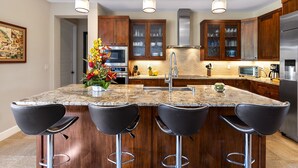 Una cocina moderna con armarios de madera, encimera de granito, electrodomésticos de acero inoxidable y taburetes negros.