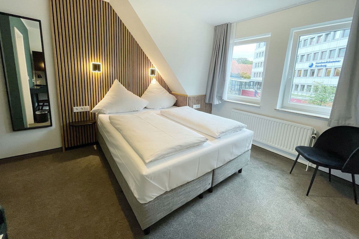 Ein Hotelzimmer mit Bett, Stuhl, Fenster und Blick auf Gebäude, sowie einer Wand mit vertikalen Linien.