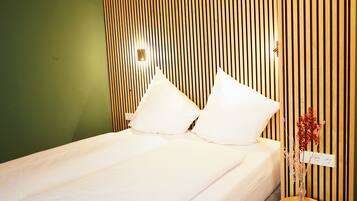 A neatly made bed with white linens and pillows, a wooden slat wall, and a small table with a vase of dried flowers.