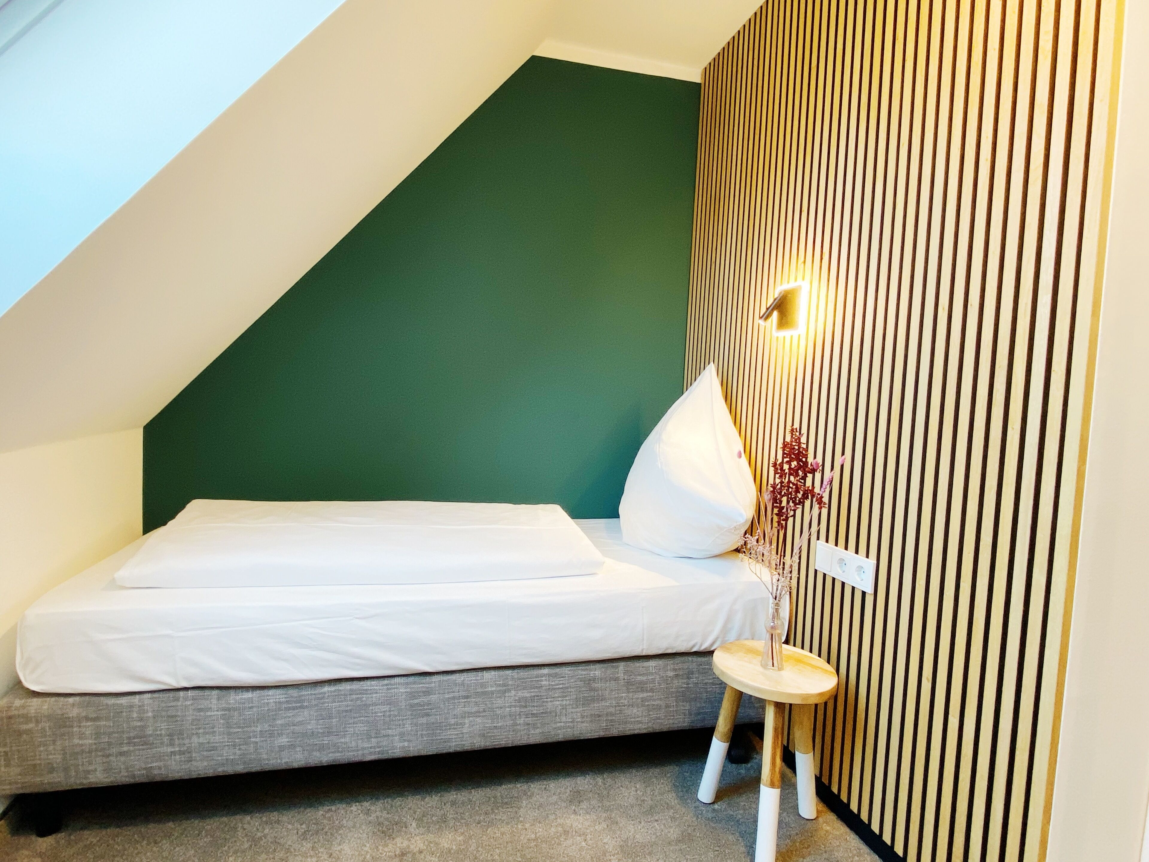 A small, modern bedroom with a sloped ceiling, a single bed, a small stool, and a wall-mounted light fixture.
