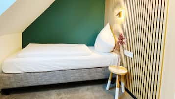 Un dormitorio pequeño y moderno con techo inclinado, una cama individual, un taburete y una lámpara fijada a la pared.