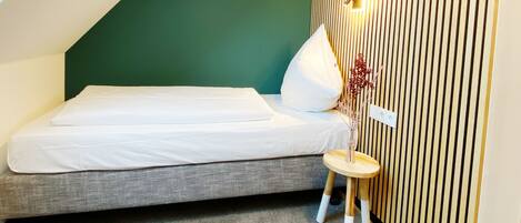 A small, modern bedroom with a sloped ceiling, a single bed, a small stool, and a wall-mounted light fixture.