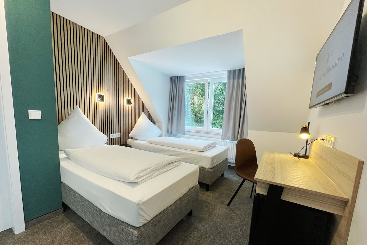 Ein Hotelzimmer mit zwei Betten, einem Schreibtisch, einem Stuhl, einem Fernseher und einem Fenster mit Vorhängen.
