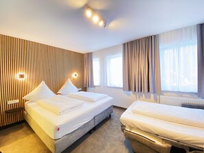 A hotel room with two beds, a wooden slat wall, a window with curtains, and a ceiling light fixture. - Hotel Xenia (Flensburg)