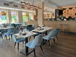 A modern restaurant interior with round tables, blue chairs, and a bar area with hexagonal light fixtures. - Hotel Xenia (Flensburg)
