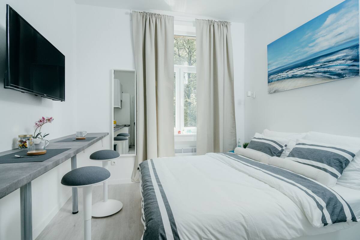 A hotel room with a bed, a desk with two stools, a TV, and a painting of the ocean.