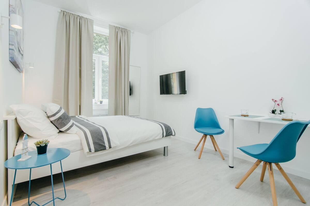 A modern bedroom with a bed, a desk, two chairs, a TV, and a window with curtains.