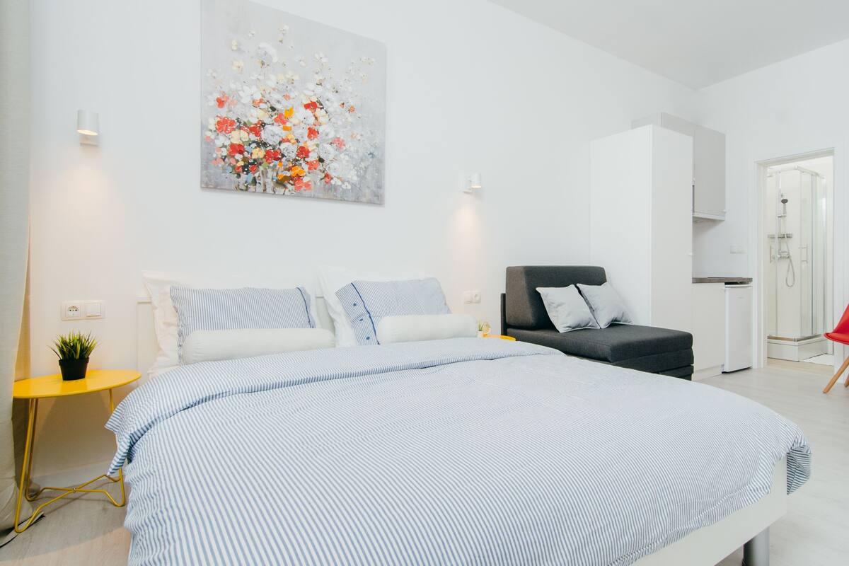 A modern bedroom with a large bed, a grey sofa, a red chair, and a small table.